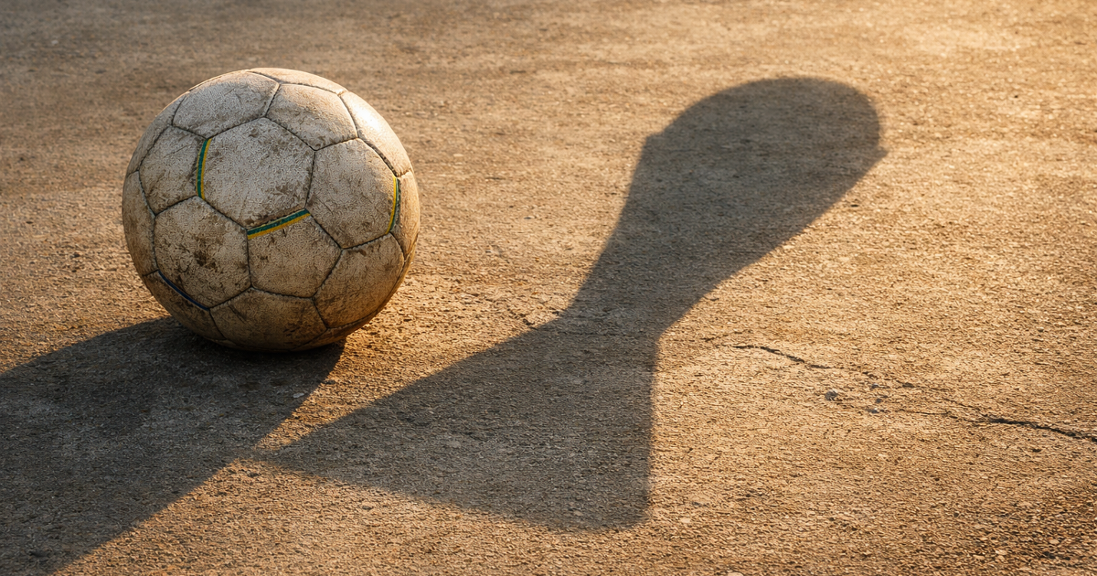 A worn football on a modest concrete pitch casts a long trophy-shaped shadow.